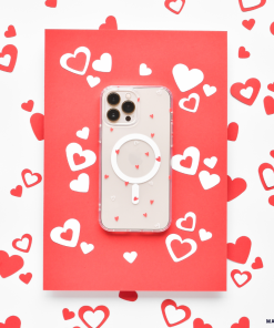 A smartphone with a high-quality Coconut Lane Clear MagSafe® Phone Case - Cute Hearts, adorned with tiny red hearts, is placed on a red card. The setup is surrounded by scattered red and white heart-shaped cutouts, creating a romantic and festive theme.