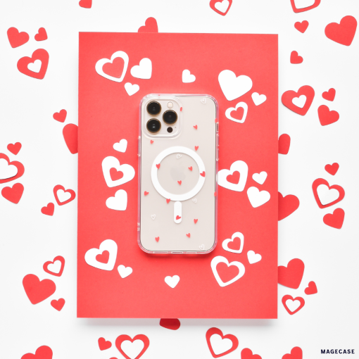 A smartphone with a high-quality Coconut Lane Clear MagSafe® Phone Case - Cute Hearts, adorned with tiny red hearts, is placed on a red card. The setup is surrounded by scattered red and white heart-shaped cutouts, creating a romantic and festive theme.