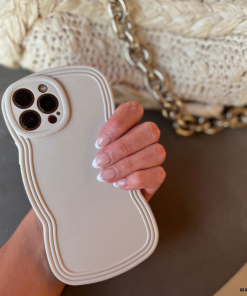 A hand with manicured nails holds a white smartphone encased in the Coconut Lane NAKD Curvy Phone Case - Vanilla, celebrated for its high protection and lightweight design. In the background, a textured beige bag adorned with a chunky gold chain strap adds an elegant touch.