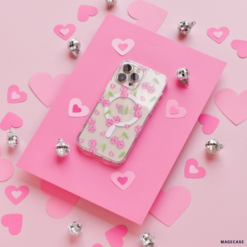 A Coconut Lane Clear MagSafe® Phone Case - Disco Cherries is placed on a hot pink rectangle surrounded by pink and light pink paper hearts. Small silver spherical decorations are scattered around, enhancing the romantic, festive vibe on a light pink background.