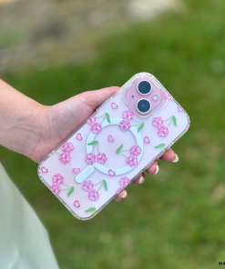 A person is holding a smartphone with Coconut Lane's Clear MagSafe® Phone Case - Disco Cherries, featuring cherry blossom designs and small hearts. The background displays a grassy area with a blurred effect.