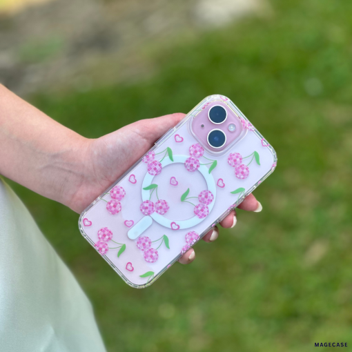 A person is holding a smartphone with Coconut Lane's Clear MagSafe® Phone Case - Disco Cherries, featuring cherry blossom designs and small hearts. The background displays a grassy area with a blurred effect.