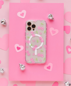 A smartphone with a Coconut Lane Clear MagSafe® Phone Case in Disco Cherries design is placed on pink paper with heart cutouts. Silver disco ball ornaments are scattered around, enhancing the playful and romantic theme.