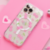 A smartphone equipped with the Coconut Lane Clear MagSafe® Phone Case - Disco Cherries, featuring pink checkered cherries, green leaves, and a white circular grip, is placed on a pink surface. Surrounding the phone are small pink heart-shaped cutouts and two tiny silver disco balls.