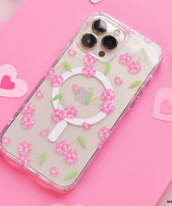 A smartphone equipped with the Coconut Lane Clear MagSafe® Phone Case - Disco Cherries, featuring pink checkered cherries, green leaves, and a white circular grip, is placed on a pink surface. Surrounding the phone are small pink heart-shaped cutouts and two tiny silver disco balls.