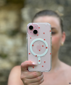 A person holds a pink iPhone covered with the Clear MagSafe® Phone Case - Cute Hearts by Coconut Lane. Small red hearts are scattered across the case, providing both high-quality protection and an aesthetic touch. The MagSafe® technology circle is visible through the case. The background is blurry, and the person is slightly out of focus, bringing attention to the phone.