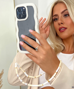 A blonde woman with long hair takes a mirror selfie using an iPhone 15 in a Curvy Phone Case - Cream by Coconut Lane, holding the phone with her left hand. She wears a light-colored jacket over a white top, showcasing a prominent ring on her ring finger and a decorative beaded phone strap.