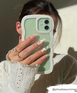 A person with dark hair, wearing a white knitted sweater, holds a green Coconut Lane Curvy Phone Case - Cream partially covering their face. Their nails are long with colorful daisy designs. The Instagram handle 
