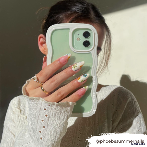 A person with dark hair, wearing a white knitted sweater, holds a green Coconut Lane Curvy Phone Case - Cream partially covering their face. Their nails are long with colorful daisy designs. The Instagram handle "@phoebesummernails" is visible in the image.