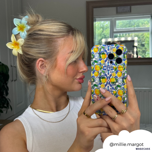 A blonde woman takes a mirror selfie with her phone adorned in the Wavy Phone Case - Amalfi Lemon by Coconut Lane. She wears a white top, gold hoop earrings, and necklaces. Her hair is tied up with large floral hair clips. The background includes a window and home interior. Instagram handle @millie.margot.