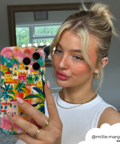 A young woman with blonde hair tied in a high bun takes a mirror selfie holding a phone adorned with the Portofino Dreams Wavy Phone Case by Coconut Lane. She is wearing a white sleeveless top, hoop earrings, and a gold necklace. Her Instagram handle 