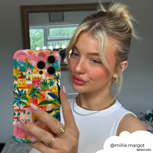 A young woman with blonde hair tied in a high bun takes a mirror selfie holding a phone adorned with the Portofino Dreams Wavy Phone Case by Coconut Lane. She is wearing a white sleeveless top, hoop earrings, and a gold necklace. Her Instagram handle "@millie.margot" is included in the bottom right corner.