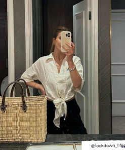 A person taking a mirror selfie in a bathroom, proudly showcasing their Curvy Phone Case - Cream from Coconut Lane on their iPhone 15. They are dressed in a white shirt tied at the waist and black pants, with a large woven straw bag resting on the counter. The Instagram handle @lockdown_lifestyle is visible in the bottom right corner.