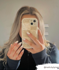 A person with long, wavy blonde hair takes a mirror selfie, covering their face with a phone adorned in Coconut Lane's Cosy Teddy Phone Case in Vanilla Biscuit. They are wearing a black top, and their nails are painted white. The handle 