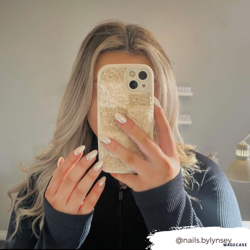 A person with long, wavy blonde hair takes a mirror selfie, covering their face with a phone adorned in Coconut Lane's Cosy Teddy Phone Case in Vanilla Biscuit. They are wearing a black top, and their nails are painted white. The handle "@nails.bylynsey" is displayed in the bottom right corner.