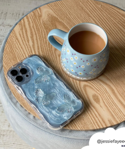 A blue mug with floral designs filled with tea sits on a round wooden table. Beside it is an iPhone encased in the Shimmer Blue Butterflies Phone Case by Coconut Lane. A white circular tag in the bottom right corner shows the handle @jessiefayeeee.