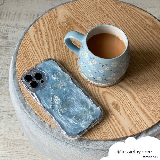 A blue mug with floral designs filled with tea sits on a round wooden table. Beside it is an iPhone encased in the Shimmer Blue Butterflies Phone Case by Coconut Lane. A white circular tag in the bottom right corner shows the handle @jessiefayeeee.