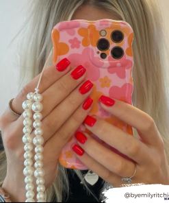 A person with blonde hair is holding a smartphone in front of their face, which is protected by Coconut Lane's Wavy Phone Case - Pink & Orange Bloom. The case features a floral design with pink and orange flowers and a string of pearls. The person has bright red nail polish. The Instagram handle 