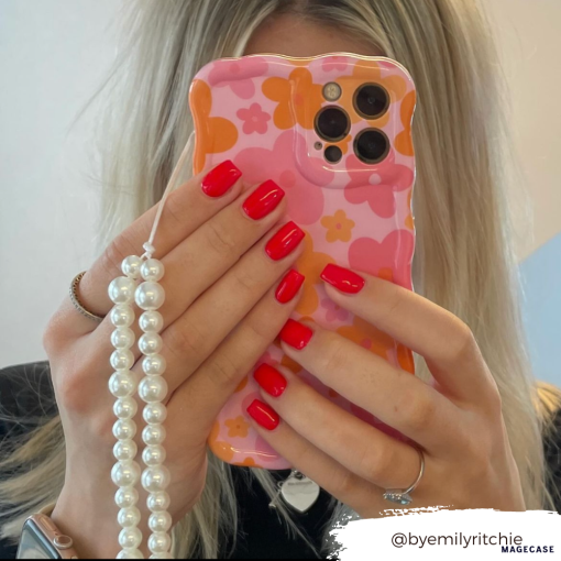 A person with blonde hair is holding a smartphone in front of their face, which is protected by Coconut Lane's Wavy Phone Case - Pink & Orange Bloom. The case features a floral design with pink and orange flowers and a string of pearls. The person has bright red nail polish. The Instagram handle "@byemilyritchie" is shown in the bottom right corner.