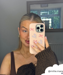 A woman with blonde hair, dressed in a brown outfit, takes a mirror selfie with her iPhone15 encased in the Coconut Lane Pearly Hearts Phone Case - Pink, featuring heart and x designs. She stands in a room where the white wall and window reflect greenery. The photo is shared via the handle @millie.margot.