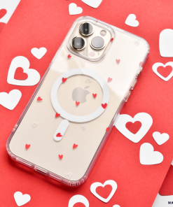 Placed on a red and white background with scattered heart-shaped cutouts, the Clear MagSafe® Phone Case - Cute Hearts by Coconut Lane offers high-quality protection while showcasing the phone's camera lenses, MagSafe® technology, and Apple logo through its transparent cover adorned with small red hearts.