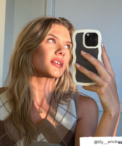 A person with long blonde hair takes a selfie in a well-lit room using a Curvy Phone Case - Cream from Coconut Lane. They are wearing a brown and white argyle sweater vest, gazing slightly upward. The Instagram handle 