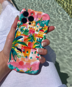 A person holding an iPhone with a colorful, tropical-themed Coconut Lane Wavy Phone Case - Portofino Dreams featuring palm trees and vibrant buildings. The person is wearing a white garment, and the background includes shimmering turquoise pool water and part of a white poolside lounger.