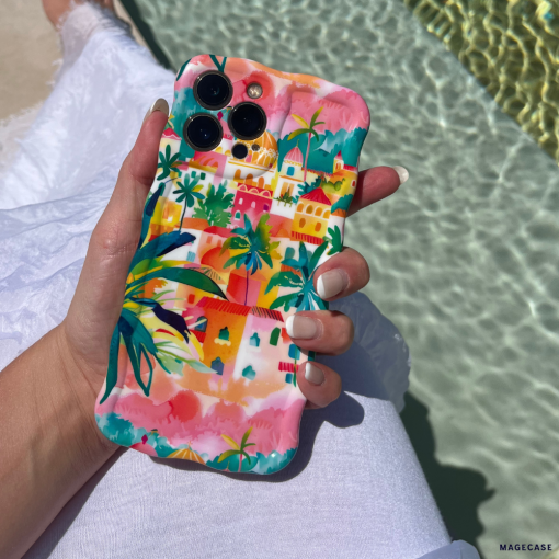 A person holding an iPhone with a colorful, tropical-themed Coconut Lane Wavy Phone Case - Portofino Dreams featuring palm trees and vibrant buildings. The person is wearing a white garment, and the background includes shimmering turquoise pool water and part of a white poolside lounger.