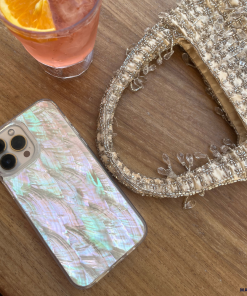 A Real Natural Shell Phone Case - White by Coconut Lane rests on a wooden table beside a textured handbag adorned with intricate beadwork. Nearby, there's a glass holding a beverage garnished with an orange slice.