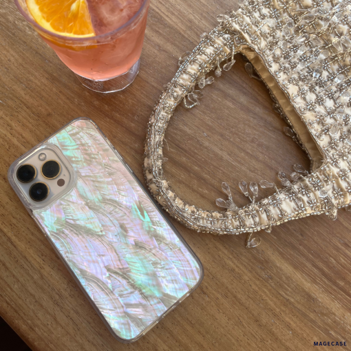 A Real Natural Shell Phone Case - White by Coconut Lane rests on a wooden table beside a textured handbag adorned with intricate beadwork. Nearby, there's a glass holding a beverage garnished with an orange slice.