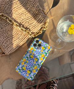 A woven straw handbag with a chunky gold chain strap is placed on a wooden table. Next to it is a phone with Coconut Lane's Wavy Phone Case in Amalfi Lemon. A glass with a clear beverage and a yellow flower floating on top is nearby. The table rests on a glass surface.