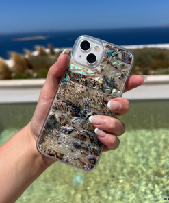 A hand with manicured nails holds a smartphone encased in the Coconut Lane Real Natural Shell Phone Case - Black, showcasing an iridescent abalone pattern. The background features a tranquil ocean view, a clear blue sky, and distant islands, evoking a serene coastal or beach setting.