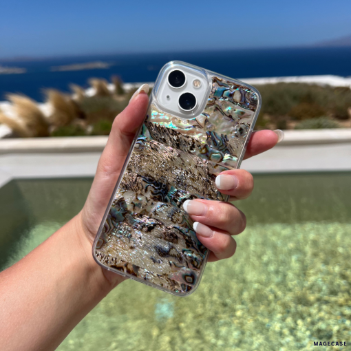 A hand with manicured nails holds a smartphone encased in the Coconut Lane Real Natural Shell Phone Case - Black, showcasing an iridescent abalone pattern. The background features a tranquil ocean view, a clear blue sky, and distant islands, evoking a serene coastal or beach setting.