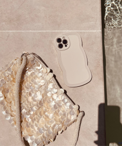 A beige smartphone with a wavy-edged NAKD Curvy Phone Case - Vanilla by Coconut Lane lies next to a shimmery bag adorned with large, circular sequins. Both lightweight items are on a light-colored surface near a pool, with ripples in the water reflecting sunlight. A shadow is partially visible in the bottom right corner.