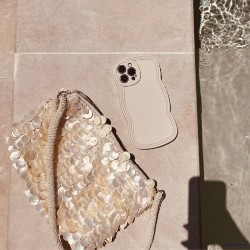 A beige smartphone with a wavy-edged NAKD Curvy Phone Case - Vanilla by Coconut Lane lies next to a shimmery bag adorned with large, circular sequins. Both lightweight items are on a light-colored surface near a pool, with ripples in the water reflecting sunlight. A shadow is partially visible in the bottom right corner.
