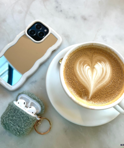 A cup of latte with heart-shaped latte art sits on a saucer atop a marble surface. Next to it are an iPhone adorned with the sleek and stylish Curvy Mirror Phone Case in cream by Coconut Lane, and a keychain holding a fuzzy gray AirPods case, with the AirPods partially visible.