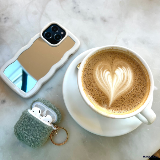 A cup of latte with heart-shaped latte art sits on a saucer atop a marble surface. Next to it are an iPhone adorned with the sleek and stylish Curvy Mirror Phone Case in cream by Coconut Lane, and a keychain holding a fuzzy gray AirPods case, with the AirPods partially visible.
