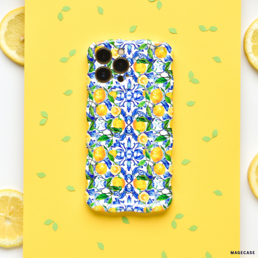 The Wavy Phone Case - Amalfi Lemon by Coconut Lane is showcased on a yellow background. Small green leaf confetti and three lemon slices are placed around it. The case boasts bright blue flowers and yellow Amalfi lemons in a vibrant pattern, providing extra protection for the smartphone.