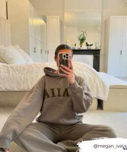 A person wearing a gray hooded sweatshirt and matching sweatpants is sitting cross-legged on a carpeted floor in front of a bed. They are taking a mirror selfie with their phone, which features the Coconut Lane Curvy Mirror Phone Case - Pink. An Instagram handle, @megan_iveyxx, is displayed in the bottom right corner.