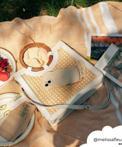 A picturesque picnic setup featuring a woven tote bag, a smartphone encased in the highly protective NAKD Curvy Phone Case - Vanilla by Coconut Lane, a plate of fruit, a small round loaf of bread, and magazines laid out on a blanket in the grass. The image is warm and sunny, depicting a relaxing outdoor scene. (@melissafleura).