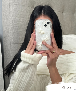 A person with long black hair wearing a white off-the-shoulder sweater holds a Coconut Lane Pearls & Bows Phone Case in front of their face, snapping a mirror selfie. A white textured headboard and white bed linen are visible in the background. A tag @_i.xa appears at the bottom right.