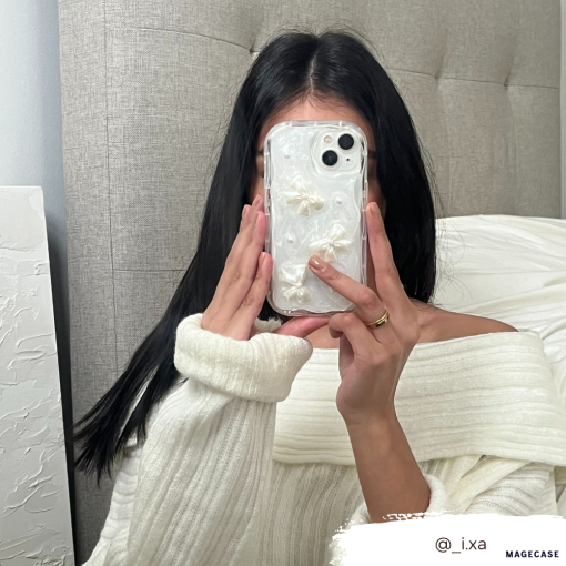 A person with long black hair wearing a white off-the-shoulder sweater holds a Coconut Lane Pearls & Bows Phone Case in front of their face, snapping a mirror selfie. A white textured headboard and white bed linen are visible in the background. A tag @_i.xa appears at the bottom right.