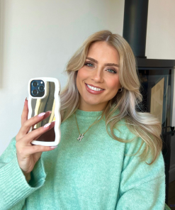 A person with long blonde hair, wearing a mint green sweater, smiles while holding up a smartphone encased in the sleek and stylish Curvy Mirror Phone Case - Cream by Coconut Lane. A necklace with a cross pendant hangs around their neck, and the background features a modern fireplace against a white wall.