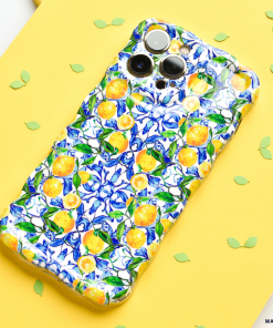 A smartphone adorned with Coconut Lane's Wavy Phone Case - Amalfi Lemon, boasting a vibrant pattern of yellow Amalfi lemons and blue flowers, is placed on a yellow surface. Green leaf-shaped confetti is scattered around the phone.