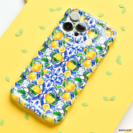 A smartphone adorned with Coconut Lane's Wavy Phone Case - Amalfi Lemon, boasting a vibrant pattern of yellow Amalfi lemons and blue flowers, is placed on a yellow surface. Green leaf-shaped confetti is scattered around the phone.