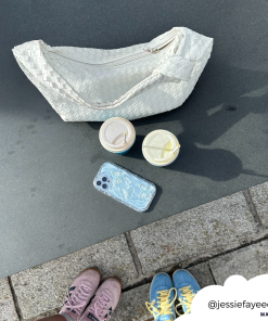 An overhead shot captures two pairs of feet—one in pink shoes, the other in blue and yellow shoes—a light-colored woven handbag, two takeaway drinks, and a smartphone encased in a Shimmer Blue Butterflies Phone Case by Coconut Lane on a stone surface. The Instagram handle @jessiefayeeee is written on the image.