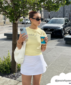 A woman is wearing a light yellow shirt and white skirt, holding a coffee cup in one hand and her phone with the Shimmer Blue Butterflies Phone Case from Coconut Lane in the other. She accessorizes with sunglasses, red earrings, and a white bag on her shoulder. She stands on a paved street with trees and cars in the background. Her Instagram handle 