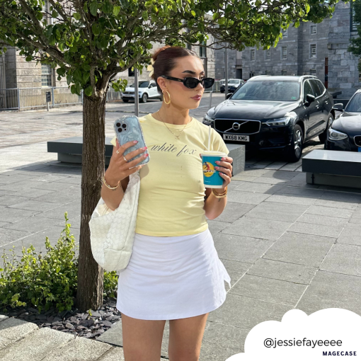A woman is wearing a light yellow shirt and white skirt, holding a coffee cup in one hand and her phone with the Shimmer Blue Butterflies Phone Case from Coconut Lane in the other. She accessorizes with sunglasses, red earrings, and a white bag on her shoulder. She stands on a paved street with trees and cars in the background. Her Instagram handle "@jessiefayeeee" is visible in the corner.