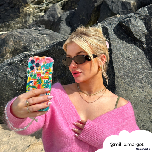 A blonde woman wearing sunglasses and a pink sweater is taking a selfie with her smartphone, which features the extra protective "Wavy Phone Case - Portofino Dreams" by Coconut Lane. She stands in front of large rocks, and her hair is tied back. Her Instagram handle "@millie.margot" is displayed in the lower right corner.