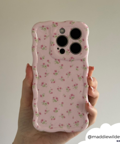A hand holding a smartphone featuring the Wavy Phone Case - Ditsy Floral Pink from Coconut Lane. The protective pink case is decorated with small, delicate floral patterns and has a distinctive wavy, textured edge that adds a unique touch. The image includes the Instagram handle 